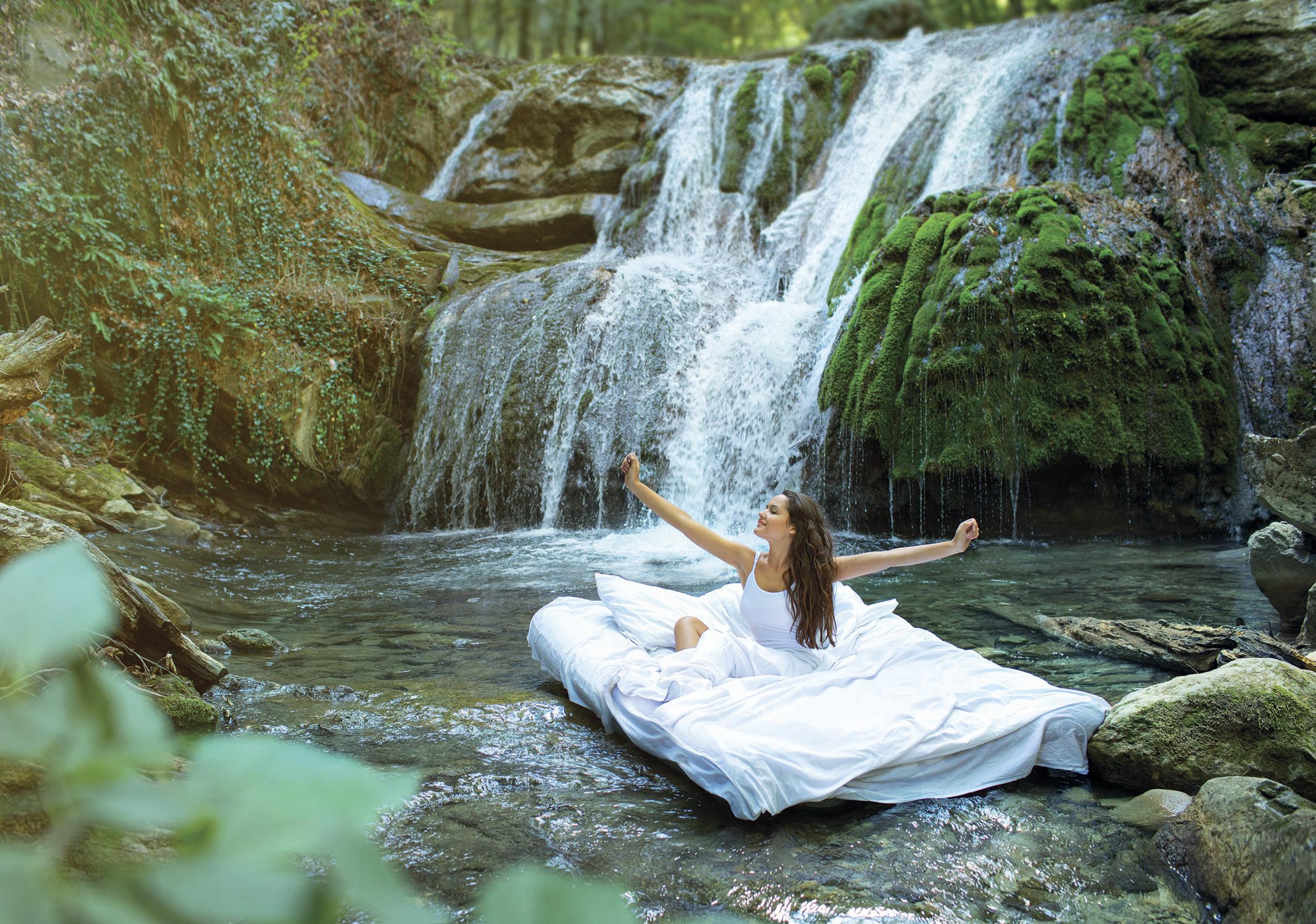 Bildkomposition Junge Frau sitzt auf einem Bett, das über einem Bach schwebt mit Wasserfall im Hintergrund
