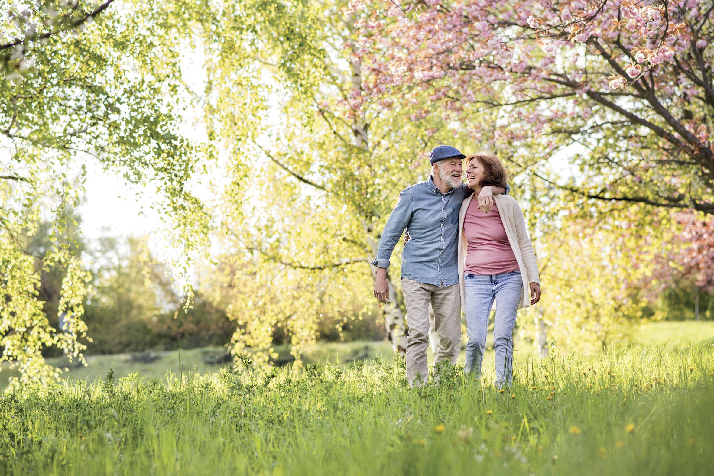 Älteres Paar geht im Frühling Arm in Arm über eine Wiese mit Bäumen